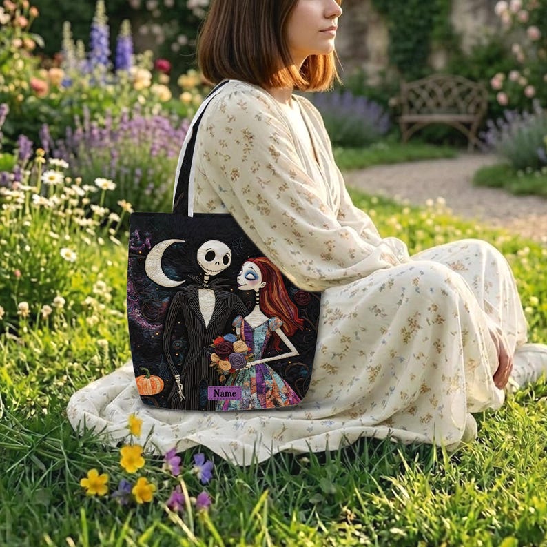 May include: A black tote bag featuring a printed illustration of a skeleton character in a pinstripe suit and a female character with long red hair holding a bouquet of flowers, with a crescent moon, a pumpkin, and a purple name tag label.