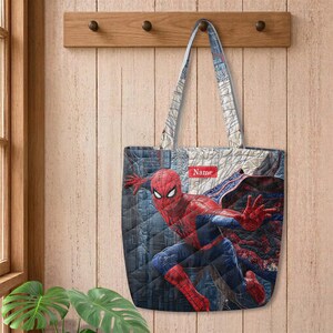 May include: A quilted tote bag featuring a graphic of the superhero Spider-Man in a dynamic pose against a dark blue cityscape background. The bag includes two shoulder straps and a small rectangular red label with the white text Name.
