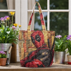 K&ouml;nnte beinhalten: Eine gesteppte Tragetasche mit einer Grafik der Figur Deadpool im rot-schwarzen Anzug vor einem gelb-orangen Stadthintergrund. Die Tasche hat zwei Schulterriemen und einen rechteckigen roten Aufn&auml;her am Boden mit dem Text Name.