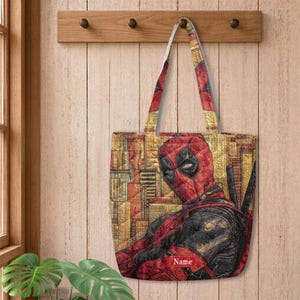 K&ouml;nnte beinhalten: Eine gesteppte Tragetasche mit einer grafischen Illustration der Figur Deadpool in einem rot-schwarzen Anzug vor einem strukturierten gold-braunen Hintergrund. Die Tasche enth&auml;lt unten ein rotes rechteckiges Etikett mit dem wei&szlig;en Text Name.