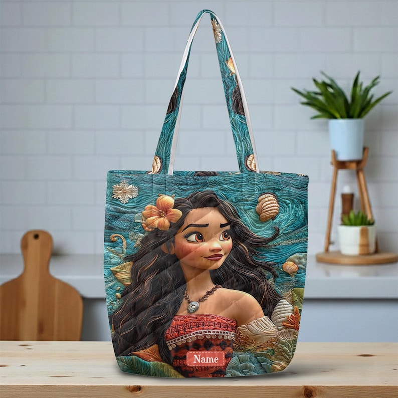 May include: A tote bag featuring a printed illustration of a character with long dark hair, a flower in their hair, and a patterned top. The background displays a textured blue ocean scene with seashells. A rectangular label with the text Name is visible.