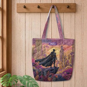 May include: A quilted tote bag featuring a graphic illustration of a black superhero figure in a cape standing on a rock overlooking a futuristic city. The design includes a purple rectangular patch with the word Name printed in white text.