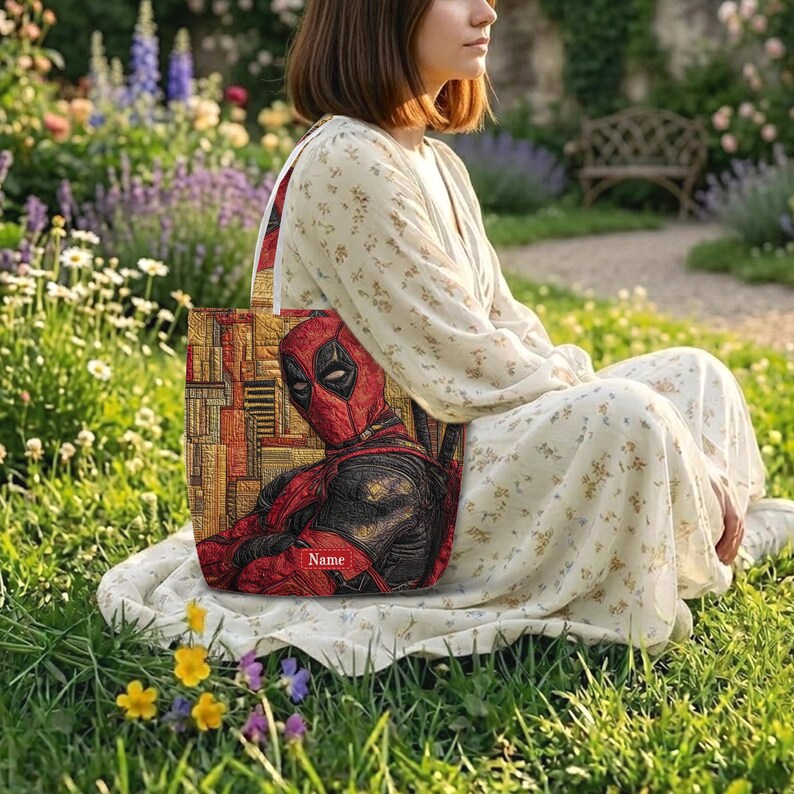 K&ouml;nnte beinhalten: Eine rechteckige Tragetasche mit einer grafischen Illustration der Figur Deadpool in Rot und Schwarz vor einem strukturierten gold-braunen Hintergrund. Die Tasche enth&auml;lt ein kleines rotes rechteckiges Etikett mit dem wei&szlig;en Aufdruck Name.