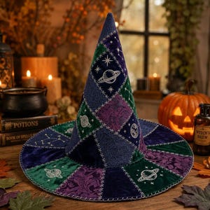 May include: A pointed hat constructed from a patchwork of velvet and textured fabric panels in shades of deep blue, purple, and green. The hat features white embroidered celestial motifs including planets, stars, and constellations.