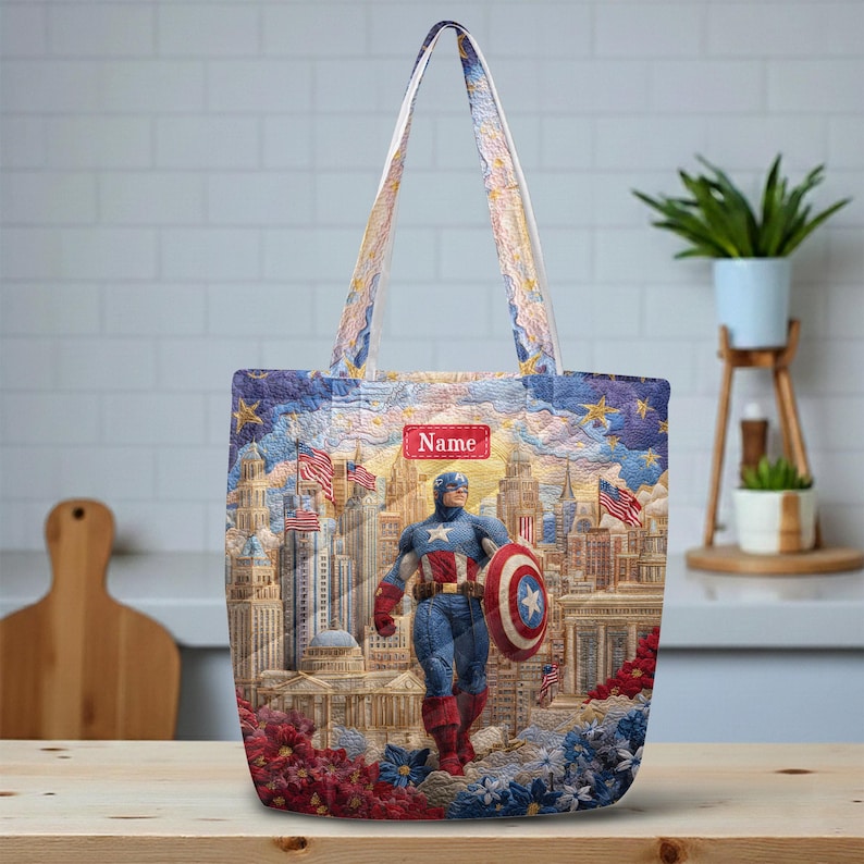 May include: A tote bag featuring a quilted-style print of Captain America standing in front of a city skyline with American flags. The design includes a red rectangular label with the text Name in the center, surrounded by blue, gold, and red floral accents.