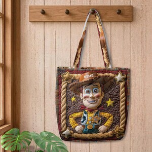 May include: A rectangular tote bag featuring a quilted brown background with a central graphic of a cowboy character wearing a brown hat with the word NAME printed on it, a yellow plaid shirt, and a blue vest, framed by a rope-like border and stars.