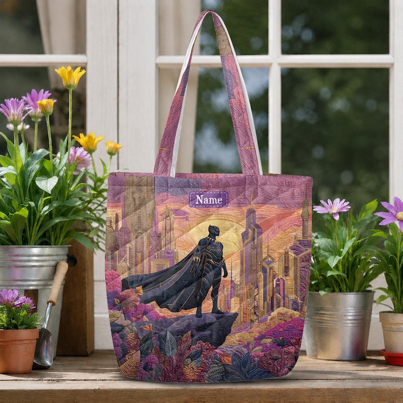 May include: A quilted tote bag featuring a graphic print of a superhero in a black suit standing on a cliff overlooking a futuristic city. The design includes purple, orange, and gold tones with a rectangular label displaying the text Name.