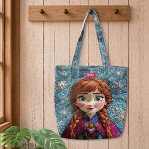 May include: A quilted tote bag featuring a blue background with white snowflake patterns and a central graphic of a character with braided auburn hair. A small pink rectangular label with the word Name is positioned above the character's head.