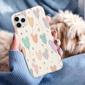 Puede incluir: Una funda de tel&eacute;fono crema con un patr&oacute;n de globos con forma de Mickey Mouse en colores pastel. Los globos son en tonos rosa, naranja, azul, verde y morado, con peque&ntilde;os corazones y estrellas.