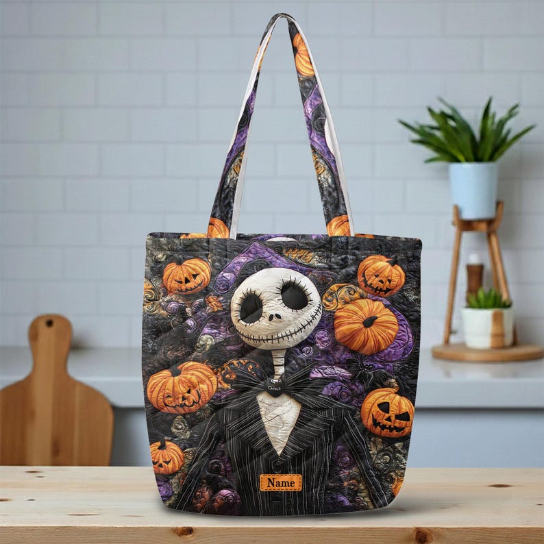 May include: A tote bag featuring a character with a white skull face and a black pinstripe suit, surrounded by orange jack-o'-lantern pumpkins on a dark purple and black patterned background. A small rectangular tag with the text Name is visible.