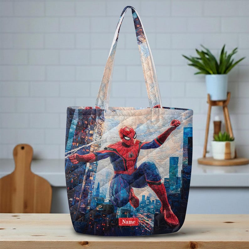 May include: A quilted tote bag featuring a graphic of Spider-Man in a dynamic pose against a city skyline background. The bag has two shoulder straps and a small rectangular red label at the bottom center with the word Name printed in white.