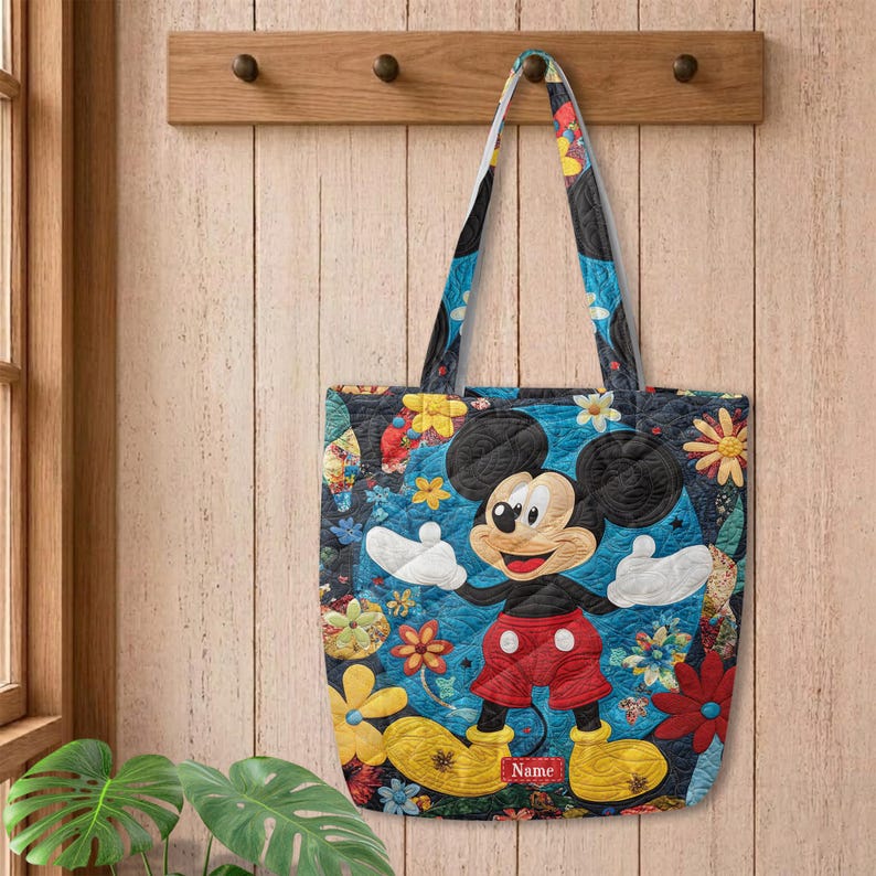May include: A quilted tote bag featuring a colourful floral pattern and a central graphic of Mickey Mouse wearing red shorts and yellow shoes. The bag includes a rectangular red name tag label at the bottom centre with the word Name printed on it.
