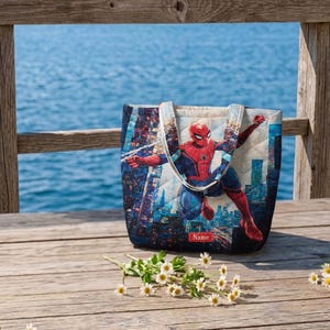 May include: A quilted tote bag featuring a graphic print of the superhero Spider-Man in a dynamic pose against a city skyline. The bag includes two shoulder straps and a small rectangular red label at the bottom center with the text Name.