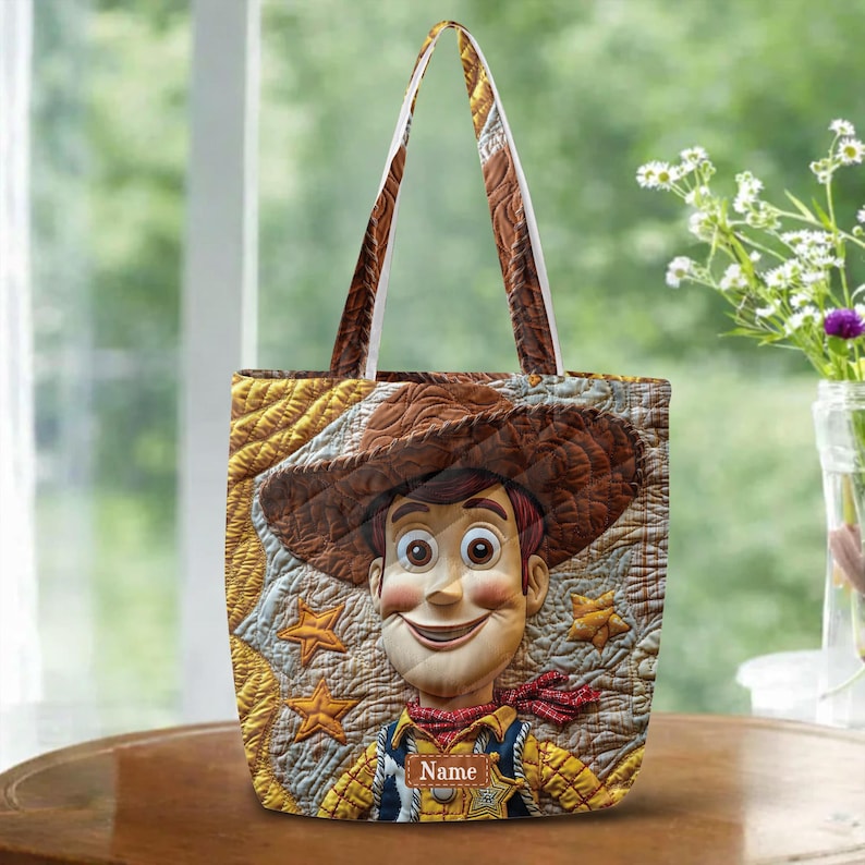 May include: A quilted tote bag featuring a graphic of a cowboy character wearing a brown hat and a yellow shirt with a red neckerchief. The background is a textured gold fabric with star patterns and a rectangular label with the text Name.