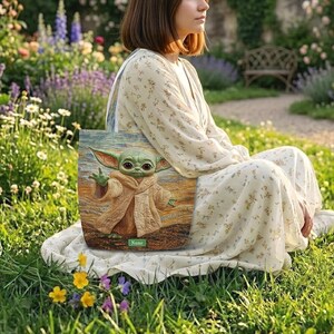 Puede incluir: Un bolso tote rectangular con una ilustraci&oacute;n impresa de una criatura verde de orejas grandes con una t&uacute;nica color canela. El bolso tiene un fondo texturizado con pinceladas arremolinadas y una etiqueta rectangular verde con la palabra Name.