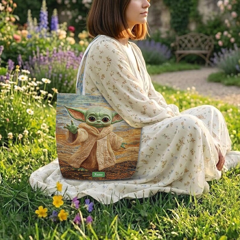 Puede incluir: Un bolso tote rectangular con una ilustraci&oacute;n impresa de una criatura verde de orejas grandes con una t&uacute;nica color canela. El bolso tiene un fondo texturizado con pinceladas arremolinadas y una etiqueta rectangular verde con la palabra Name.