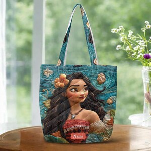 May include: A tote bag featuring a printed illustration of a character with long dark hair, wearing a patterned top and a necklace. The background displays a textured blue ocean scene with scattered seashells and an orange flower. The word Name is printed.