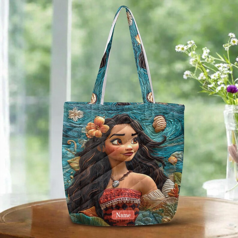 May include: A tote bag featuring a printed illustration of a character with long dark hair, wearing a patterned top and a necklace. The background displays a textured blue ocean scene with scattered seashells and an orange flower. The word Name is printed.