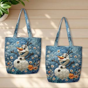 May include: Two quilted tote bags featuring a blue background with a printed snowman character, orange carrot nose, and white snowflake and flower patterns. Each bag includes a rectangular label with the text Name at the top center.