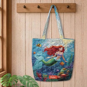 Puede incluir: Un bolso tote acolchado con una vibrante escena submarina que muestra una sirena con largo cabello rojo, cola verde y un top de conchas moradas. El dise&ntilde;o incluye corales coloridos, peces peque&ntilde;os y una etiqueta roja con el texto Name.