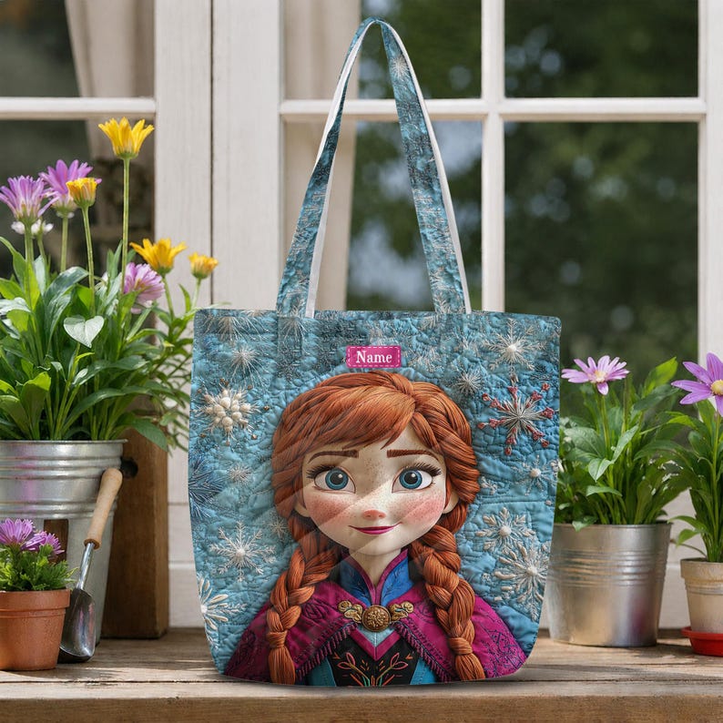 May include: A quilted tote bag featuring a graphic of a character with red braided hair and a blue and purple outfit. The background is a light blue fabric with a white snowflake pattern. A small pink rectangular label displays the word Name.