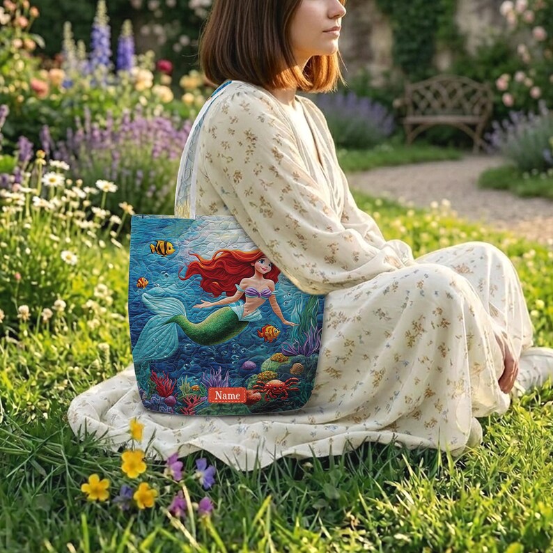 Puede incluir: Un bolso tote con una vibrante escena submarina que muestra una sirena de pelo rojo, corales coloridos y peces tropicales. El bolso incluye una etiqueta rectangular roja en la parte inferior central con la palabra Name impresa en blanco.