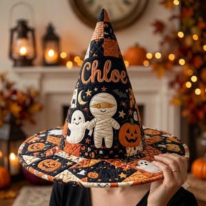 May include: A quilted witch hat featuring a black and orange patchwork pattern with embroidered Halloween motifs including a mummy, a ghost, a pumpkin, and a black cat. The name Chloe is embroidered in orange script on the front of the hat.