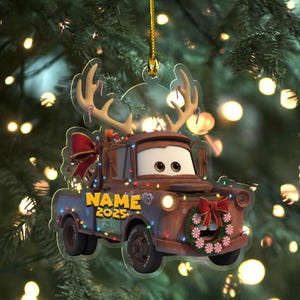 May include: A clear acrylic Christmas ornament shaped like the character Mater from the movie Cars. The ornament features a brown truck with antlers, a wreath, and the text "NAME 2025". It is decorated with Christmas lights and a red bow.