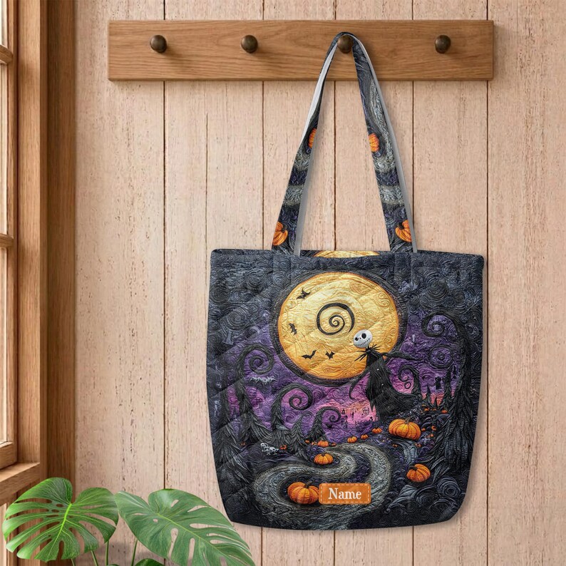 May include: A quilted tote bag featuring a dark, swirling night sky, a large yellow moon, a skeletal figure, and orange pumpkins. The design includes a rectangular patch with the word Name printed in white text on a brown background.