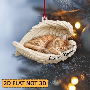 May include: A flat, two-dimensional hanging ornament featuring a ginger cat sleeping inside a pair of cream-colored angel wings. The ornament includes a red string for hanging and the black cursive text Custom Name printed on the lower right side.