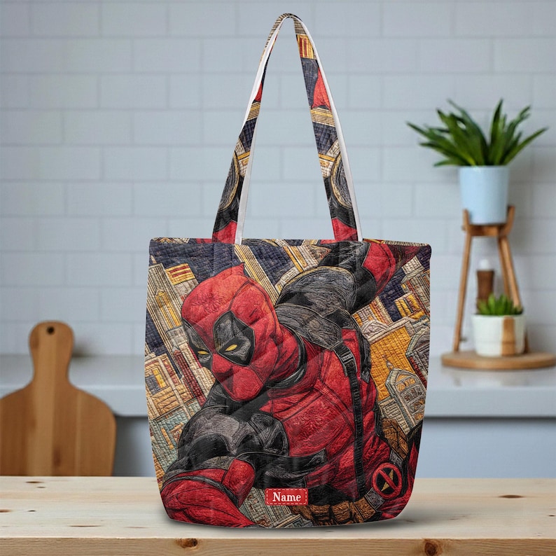 以下が含まれることがあります： 赤と黒のスーツを着たデッドプールのキャラクターが都市の背景に描かれたグラフィックイラスト入りのキルティングトートバッグ。バッグには2本のショルダーストラップがあり、下部にはNameという文字が入った小さな赤い長方形のラベルが付いています。