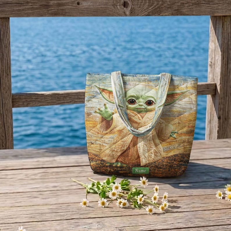 Puede incluir: Un bolso tote rectangular con una ilustraci&oacute;n impresa de una criatura verde de orejas grandes con una t&uacute;nica color canela. El bolso tiene dos asas de tela y una peque&ntilde;a etiqueta rectangular verde con la palabra Name impresa en blanco.