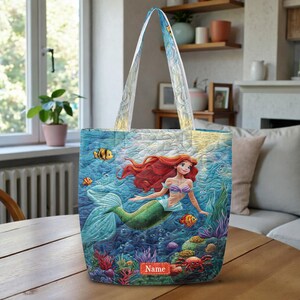 Puede incluir: Un bolso tote acolchado con una vibrante escena submarina que muestra a una sirena con largo cabello rojo, cola verde y top de conchas moradas. El dise&ntilde;o incluye corales coloridos, peque&ntilde;os peces tropicales y un cangrejo. Una etiqueta roja rectangular dice Name.