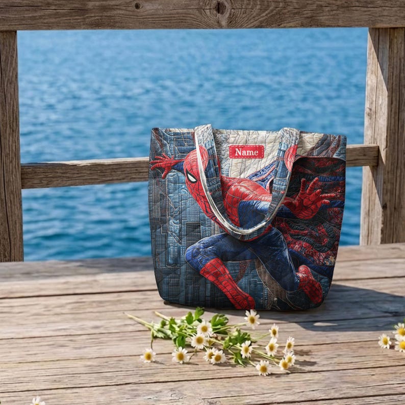 May include: A quilted tote bag featuring a graphic print of the superhero Spider-Man in a dynamic pose against a city skyline background. The bag includes a rectangular red patch near the top with the white text Name printed on it.