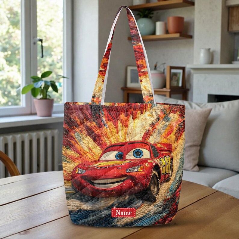 May include: A quilted tote bag featuring a vibrant, textured illustration of a red race car with blue eyes and a wide smile. The background displays a dynamic, colorful explosion of orange, yellow, and blue streaks. A small red rectangular label with the text Name is centered at the bottom.