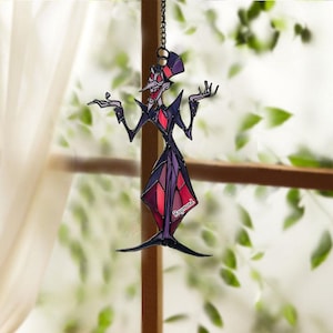 May include: A stained glass suncatcher depicting a cartoon character in a purple and red outfit, with a matching hat. The character has a long nose and is posed with arms outstretched. The name "Raymond" is visible on the design.