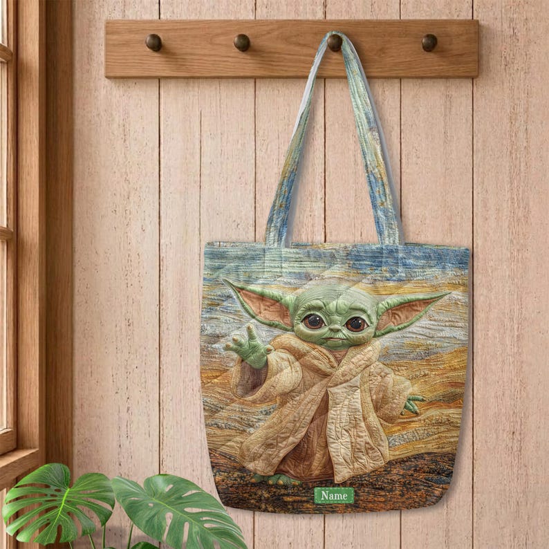 Puede incluir: Un bolso tote rectangular con la imagen impresa de una criatura verde de orejas grandes que viste una t&uacute;nica color canela. El fondo muestra un paisaje texturizado y pict&oacute;rico en tonos azules, amarillos y marrones. Se ve una peque&ntilde;a etiqueta verde con el texto Name.