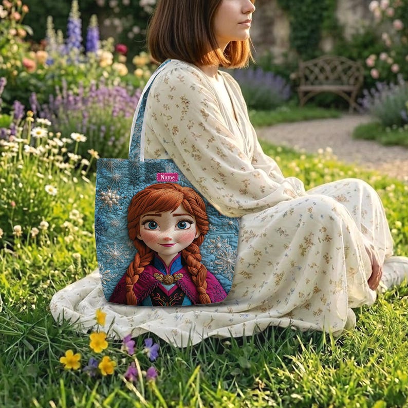 May include: A quilted tote bag featuring a printed image of a character with red braided hair, blue eyes, and a magenta cape. The bag has a blue background with white snowflake patterns and a small rectangular label with the text Name.