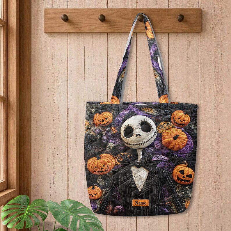 May include: A quilted tote bag featuring a graphic of a skeleton character in a pinstripe suit surrounded by orange jack-o'-lantern pumpkins and purple accents. The bag includes a rectangular label with the text Name printed on the front.
