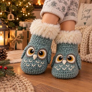 May include: A pair of teal blue crochet slippers featuring an owl face design with large yellow and black eyes, a white beak, and white V-shaped stitching. The slippers have a thick, fluffy white faux fur cuff around the top opening.