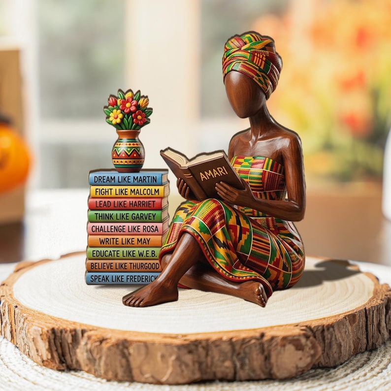 May include: A wooden figurine of a woman reading a book titled "AMARI", wearing a colorful patterned head wrap and dress. A stack of books with inspirational phrases and a small vase with flowers are next to her. The figurine is on a wooden slice.