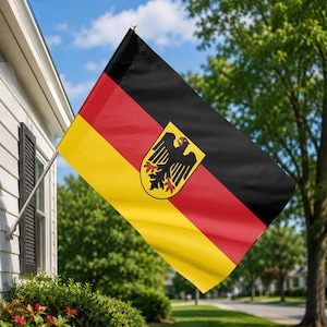 May include: A German flag with black, red, and yellow stripes, and a coat of arms featuring a black eagle on a yellow shield. The flag is displayed outdoors, mounted on a pole, with a residential backdrop.