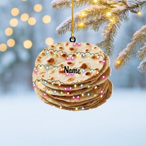 May include: A stack of flatbread Christmas ornament with a string of colorful lights. The word "Name" is written on the ornament in a cursive font. The ornament is hanging from a gold string.