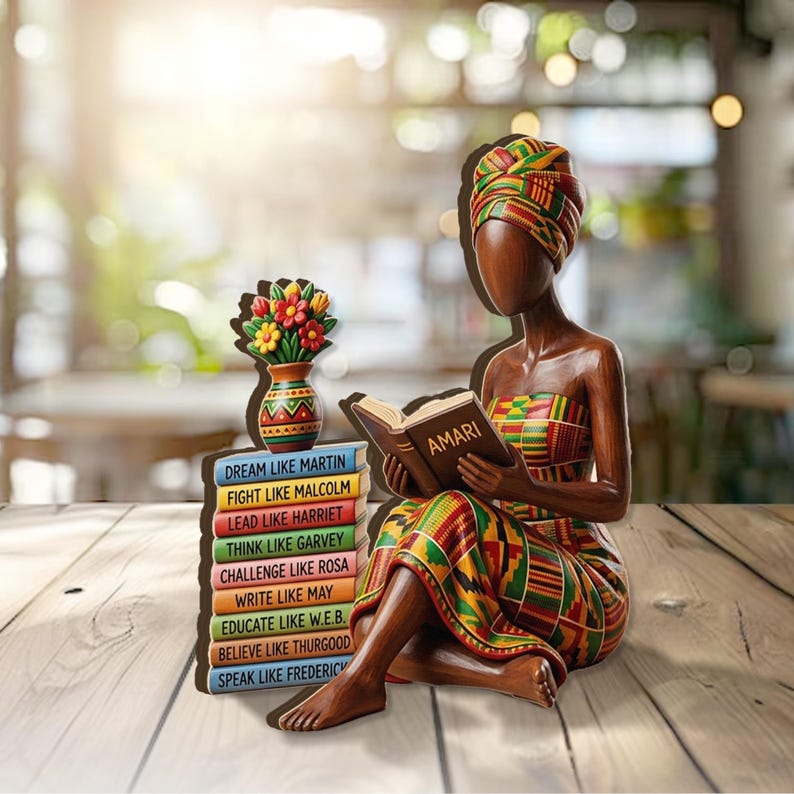 May include: A wooden figurine of a woman reading a book with the title "AMARI". She is wearing a colorful patterned dress and head wrap. Beside her is a stack of books with inspirational phrases and a vase with flowers.