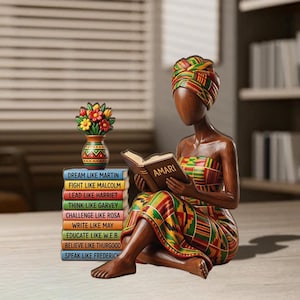 May include: A wooden figurine of a woman reading a book titled "AMARI", wearing a colorful patterned dress and head wrap. A stack of books with inspirational phrases and a vase of flowers are next to her. The figurine is brown and has a smooth finish.
