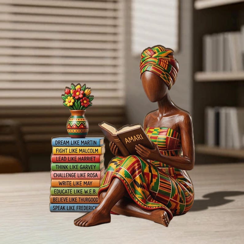 May include: A wooden figurine of a woman reading a book titled "AMARI", wearing a colorful patterned dress and head wrap. A stack of books with inspirational phrases and a vase of flowers are next to her. The figurine is brown and has a smooth finish.