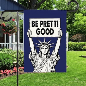 May include: A navy blue garden flag features a graphic of the Statue of Liberty holding a sign that reads "BE PRETTI GOOD" in bold white letters. The flag is displayed on a black metal stand in a garden setting.