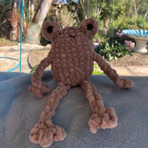 May include: A crocheted plush frog toy with a pink body and black eyes. The frog is sitting on a blue fuzzy surface.