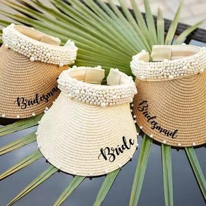 May include: Three straw visors with pearl accents. One visor is white and says "Bride" in black script. The other two are tan and say "Bridesmaid". The visors are decorated with pearls around the brim.