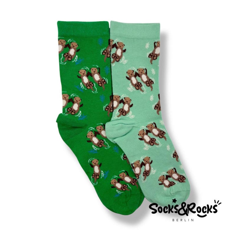 May include: Two colorful socks, one green and one teal, are displayed. Both socks feature a repeating pattern of cartoon otters swimming in water. The socks have the text "Socks&Rocks BERLIN" printed on them.